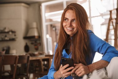 Mutfakta rahat bir şekilde otururken gülümseyen ve cep telefonu kullanan mutlu kızıl kadın görüntüsü