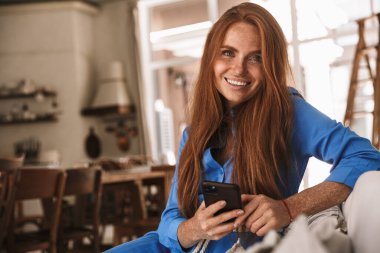 Mutfakta rahat bir şekilde otururken gülümseyen ve cep telefonu kullanan mutlu kızıl kadın görüntüsü