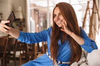 Mutfakta rahat bir şekilde otururken cep telefonunda selfie çeken mutlu kızıl kadın görüntüsü