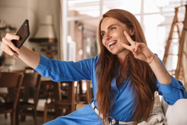 Mutlu bir kadının barış jesti yaptığı ve rahat bir mutfakta otururken cep telefonuyla selfie çektiği bir görüntü.