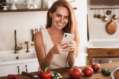 Gülümseyen genç bir kadın mutfakta durup taze salata yaparken cep telefonu kullanıyor.