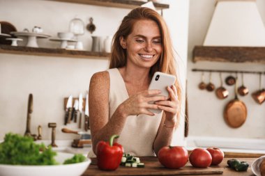 Gülümseyen genç bir kadın mutfakta durup taze salata yaparken cep telefonu kullanıyor.