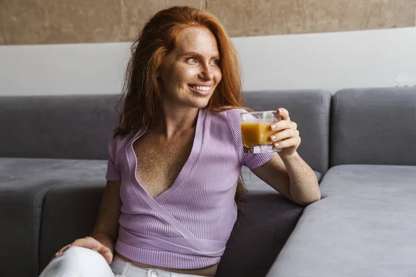 Imagen Pelirroja Alegre Mujer Sonriendo Bebiendo Jugo Mientras Está Sentada — Foto de Stock