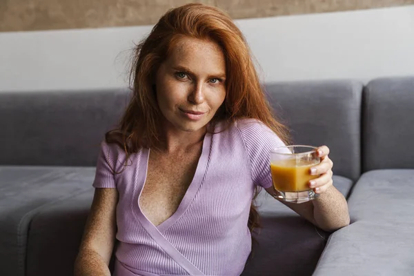 Kızıl saçlı, memnun bir kadının evde oturmuş meyve suyu içişi.