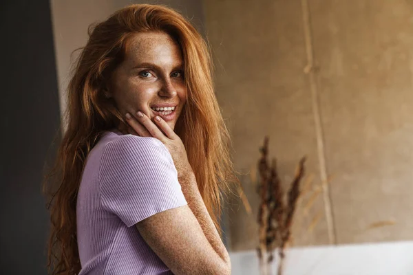 Kızıl saçlı, mutlu bir kadının oturma odasında poz verirken gülümseyen ve kameraya bakan görüntüsü