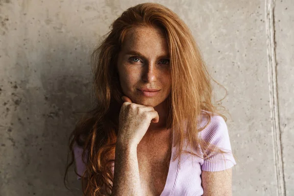 Kızıl saçlı kadının evde beton duvarın yanında poz verirken kameraya baktığı görüntü