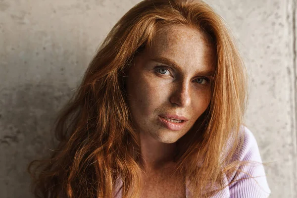 Kızıl saçlı kadının evde beton duvarın yanında poz verirken kameraya baktığı görüntü