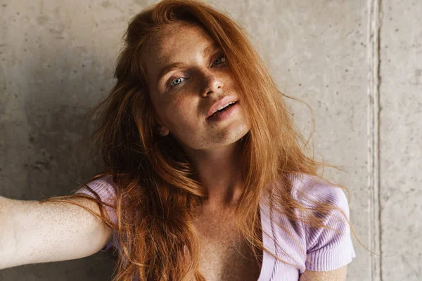 Kızıl saçlı, şehvetli bir kadının evde beton duvarın yanında poz verirken kendi fotoğrafını çekmesi.