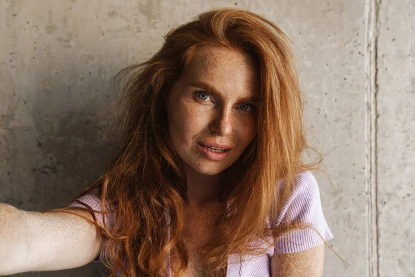 Kızıl saçlı, şehvetli bir kadının evde beton duvarın yanında poz verirken kendi fotoğrafını çekmesi.