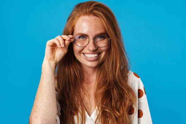 Uzun kızıl saçlı, güzel bir kadının portresi. Yazlık kıyafetler ve güneş gözlüğü takıyor. Mavi arka planda tek başına duruyor.