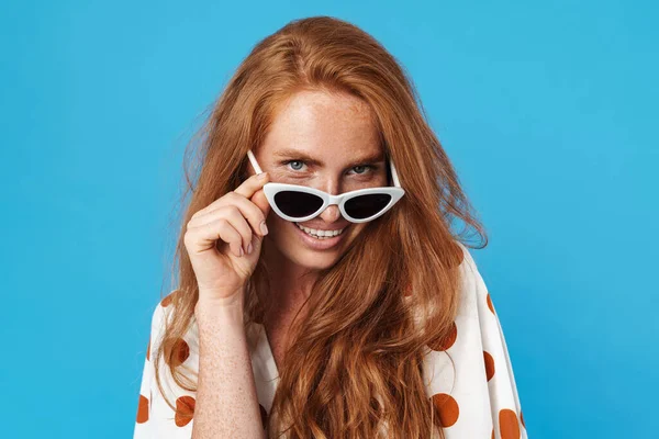 Uzun kızıl saçlı, güzel bir kadının portresi. Yazlık kıyafetler ve güneş gözlüğü takıyor. Mavi arka planda tek başına duruyor.