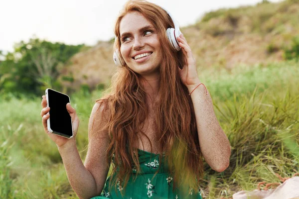 身着夏装的年轻而快乐的女人 坐在公园的绿草上 一边用智能手机和耳机听着音乐 — 图库照片