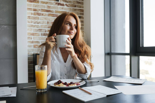 Image of pleased ginger businesswoman smiling and having breakfast in kitchen at home