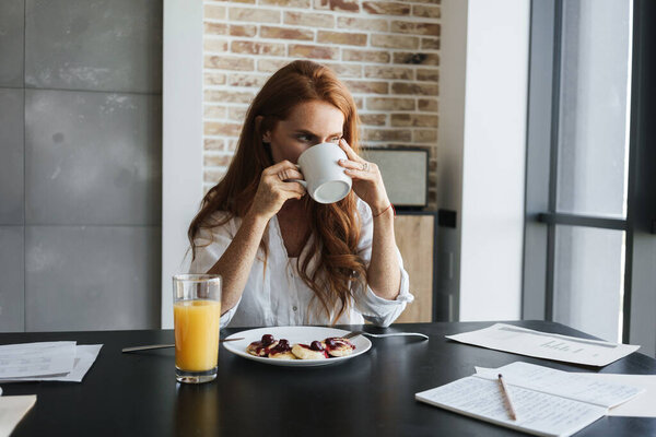 Image of young ginger businesswoman drinking coffee while having breakfast in kitchen at home