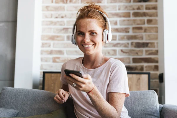 Evde kanepede otururken kulaklık ve cep telefonu kullanan gülümseyen genç bir kadın görüntüsü