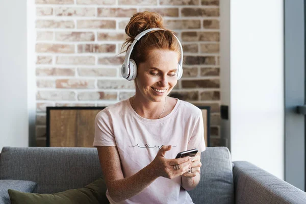 Evde kanepede otururken kulaklık ve cep telefonu kullanan gülümseyen genç bir kadın görüntüsü