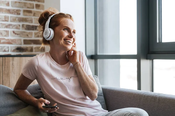 Evde kanepede otururken kulaklık ve cep telefonu kullanan gülen genç bir kadın görüntüsü