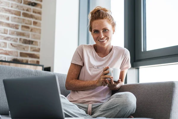 Neşeli kızıl saçlı bir kadının kahve içip dizüstü bilgisayar kullanması.