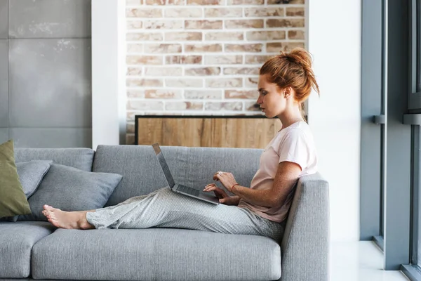 Evde kanepede otururken dizüstü bilgisayarla çalışan odaklanmış kızıl saçlı bir kadın.