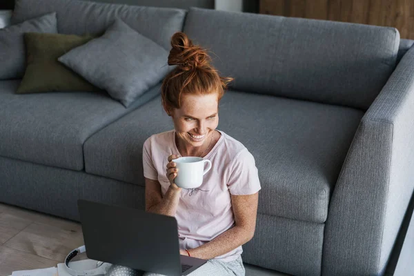 Kahvesini yudumlayan ve dizüstü bilgisayar kullanan neşeli bir kızıl kadın resmi.