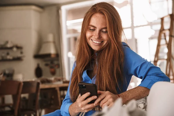 Mutfakta rahat bir şekilde otururken gülümseyen ve cep telefonu kullanan mutlu kızıl kadın görüntüsü