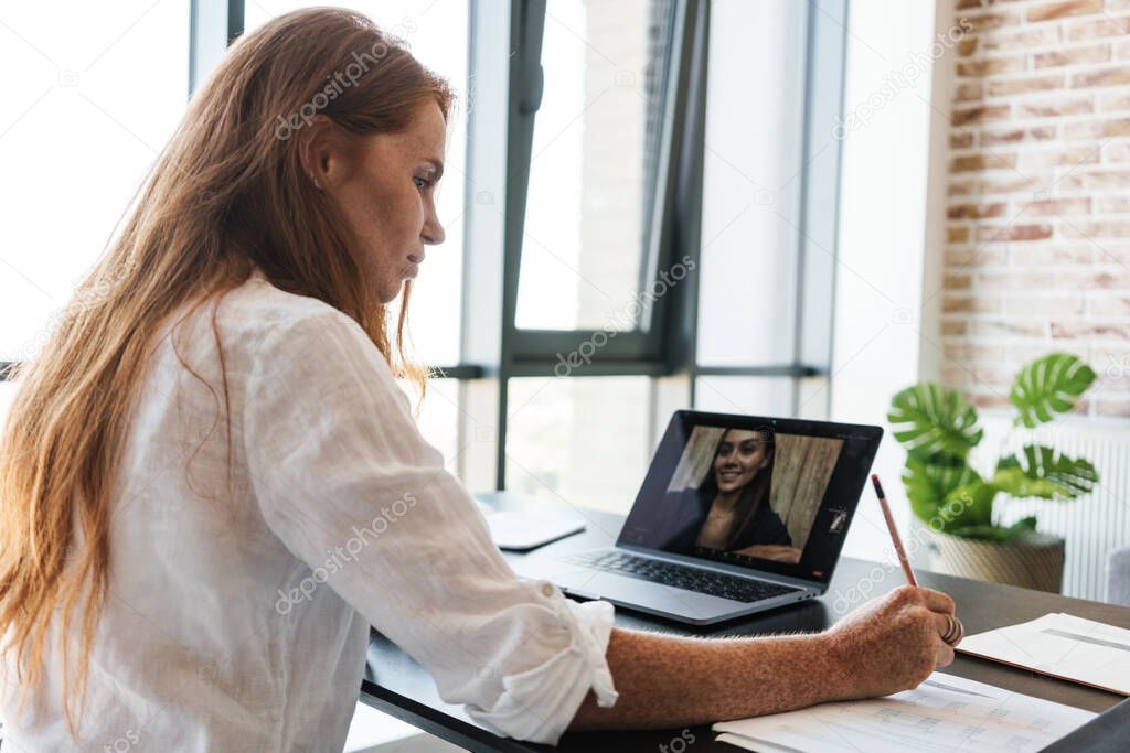 Image of redhead businesswoman making video call on laptop while sitting at table in home