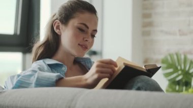 Mutlu genç bir kadın evdeki oturma odasında oturmuş kitap okuyor.