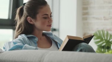 Memnun genç bir kadın evdeki oturma odasında oturmuş kitap okuyor.