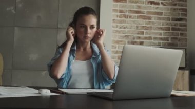 Kulaklık takan ciddi bir genç kadın laptopuyla evdeki masada oturuyor.