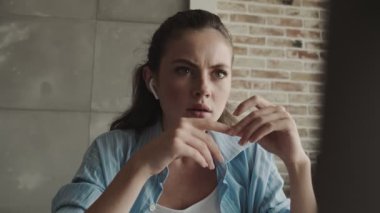 Kulaklık takan çekici genç bir kadın internette bilgisayarını kullanarak evdeki masada oturuyor.