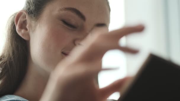 一位面带微笑的年轻女子的近照是坐在客厅里看书 — 图库视频影像