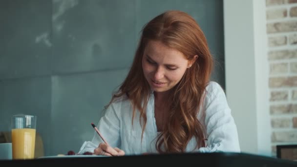 メモを書きながら家の中でテーブルに座って若い美しい赤毛の女性 — ストック動画