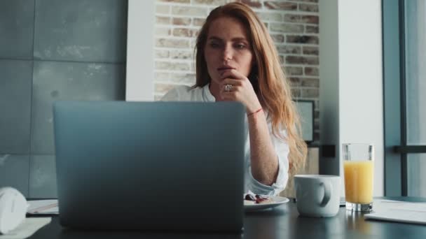 若いです深刻な赤毛の女性は ラップトップコンピュータを使用している間 屋内でテーブルに座っています — ストック動画