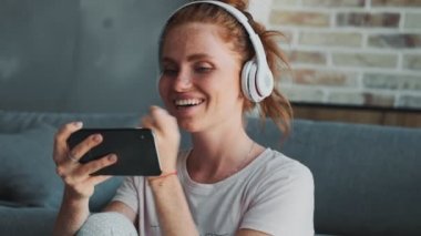 Evde genç, pozitif saçlı bir kadın cep telefonu kullanırken kulaklıkla müzik dinliyor.