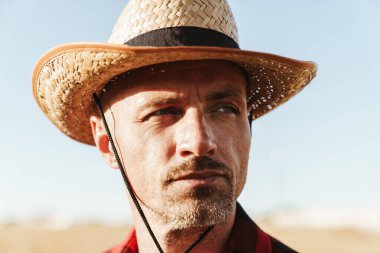 Hasır şapkalı, sakalsız yetişkin bir adam mısır gevreği tarlasında dikilirken yan bakıyor.