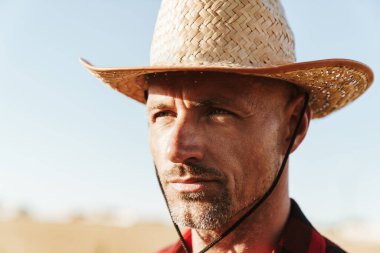 Hasır şapkalı, sakalsız yetişkin bir adam mısır gevreği tarlasında dikilirken yan bakıyor.