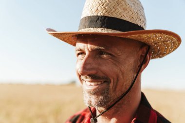 Hasır şapkalı memnun yetişkin bir adam mısır gevreği tarlasında dikilirken yan bakıyor.