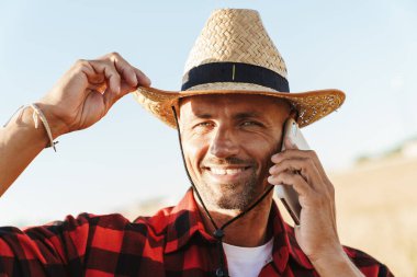 Hasır şapkalı gülümseyen yetişkin adamın görüntüsü. Mısır gevreği tarlasında dururken cep telefonuyla konuşuyor.