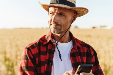 Hasır şapkalı gülümseyen yetişkin bir adamın mısır gevreği tarlasında dururken cep telefonu kullanması.