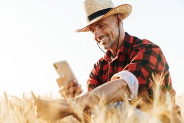 Hasır şapkalı gülümseyen yetişkin bir adamın mısır gevreği tarlasında cep telefonuyla fotoğraf çekmesi.