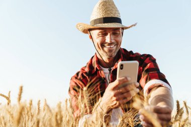 Hasır şapkalı gülümseyen yetişkin bir adamın mısır gevreği tarlasında cep telefonuyla fotoğraf çekmesi.