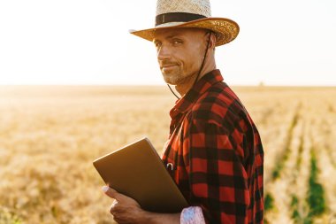 Hasır şapkalı, elinde dizüstü bilgisayarla mısır gevreği tarlasında duran memnun yetişkin bir adam görüntüsü