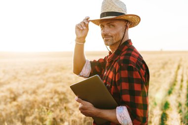 Hasır şapkalı, elinde dizüstü bilgisayarla mısır gevreği tarlasında duran memnun yetişkin bir adam görüntüsü