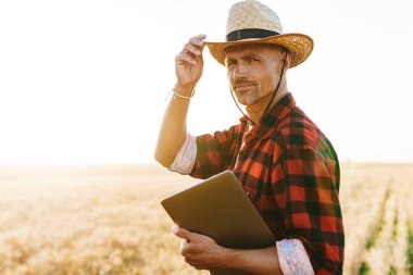 Hasır şapkalı, elinde dizüstü bilgisayarla mısır gevreği tarlasında duran memnun yetişkin bir adam görüntüsü