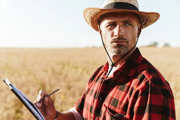 Hasır şapkalı, tahıl gevreği tarlasında durmuş kağıtlarla çalışan, traşsız bir yetişkin resmi.