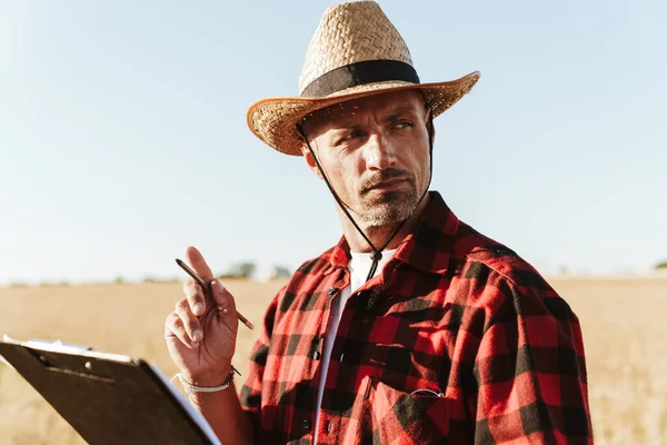 Hasır şapkalı, tahıl gevreği tarlasında durmuş kağıtlarla çalışan, traşsız bir yetişkin resmi.