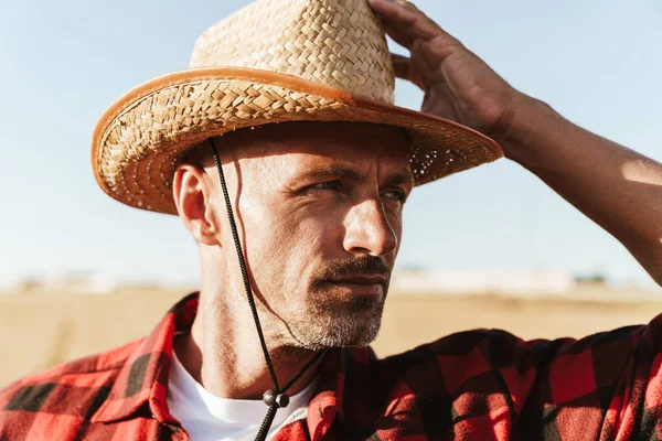 Hasır şapkalı, sakalsız yetişkin bir adam mısır gevreği tarlasında dikilirken yan bakıyor.