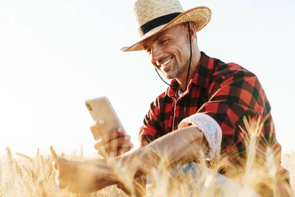 Hasır şapkalı gülümseyen yetişkin bir adamın mısır gevreği tarlasında cep telefonuyla fotoğraf çekmesi.