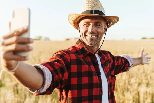 Hasır şapkalı, mutlu yetişkin bir adamın resmi. Mısır gevreği tarlasında gülümseyen ve cep telefonundan selfie çeken.