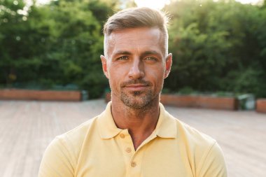 Sarı tişörtlü, kendinden emin orta yaşlı bir adamın yaz parkında yürürken kameraya baktığı görüntüler.